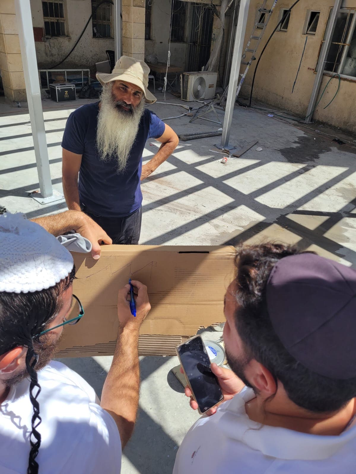 סקירת תוכנית על קרטון באתר — קבלן בכובע סוכך לצד שני אנשי קהילה צעירים