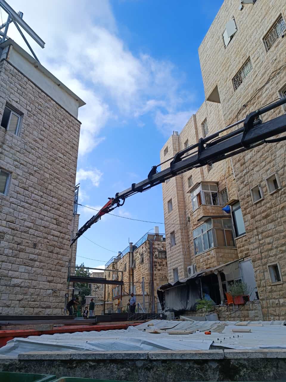 זרוע מנוף משאית מפרקי (knuckle-boom) משתרעת מעל מבני המגורים — שלב ההנפה בקריית אמשינוב